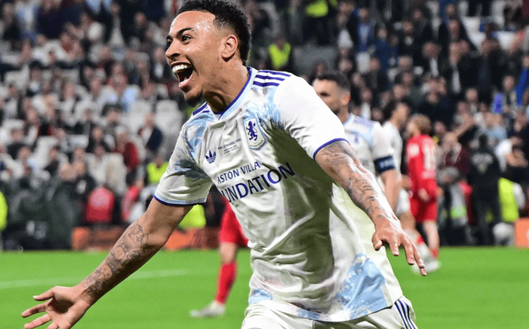Footballer in a white kit with blue trim celebrates a goal on the pitch with arms outstretched as fans cheer in the background.