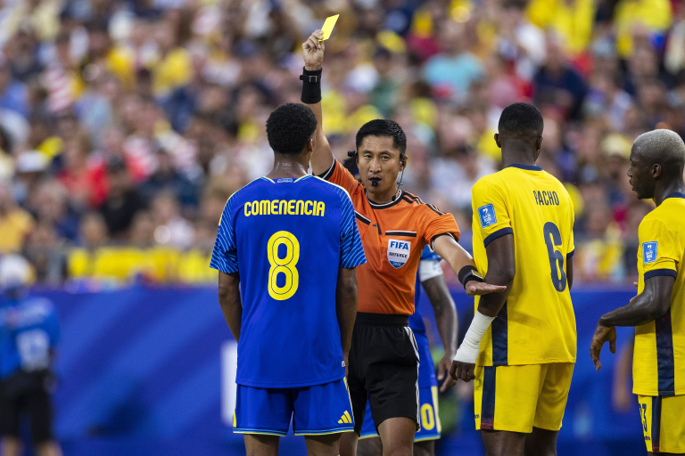 Soccer referee raises a yellow card to players during a match, with players in blue and yellow jerseys nearby.