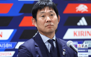 Asian man in a navy pinstripe suit speaking at a press conference, microphone in front, colorful sponsor backdrop behind.