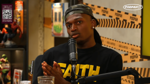 Podcast host wearing a cap speaks into a large microphone during a radio interview, with Podpah branding in the background.