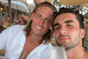 Two friends take a sunny selfie at an outdoor beachside restaurant, each holding a glass of beverage and smiling at the camera.