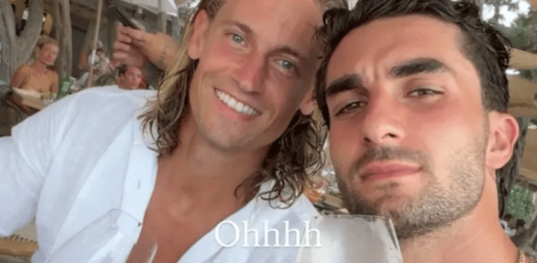 Two men smiling at the camera in a sunny outdoor restaurant, with wine glasses in view.",