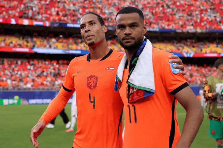 Two Dutch soccer players in orange jerseys pose together on the field with a cheering crowd behind them.