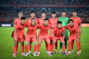 Eleven soccer players in orange uniforms pose for a team photo on the grass field, with a goalkeeper in green in the back row and stadium seating behind them.