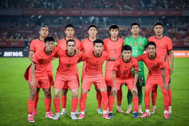 Eleven soccer players in orange uniforms pose for a team photo on the grass field, with a goalkeeper in green in the back row and stadium seating behind them.