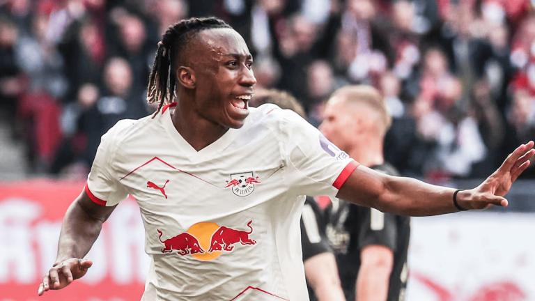 Soccer player in a white RB Leipzig jersey celebrating with arms outstretched on the pitch, crowd behind him.