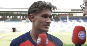 Close-up of a male footballer in a blue-red jersey speaking into a red microphone with a Bayern Munich crest; stadium stands blurred in the background.