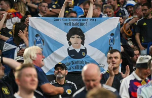 Crowd of football fans at a match holding a Scotland flag banner with a central caricature of a player and text along the top.