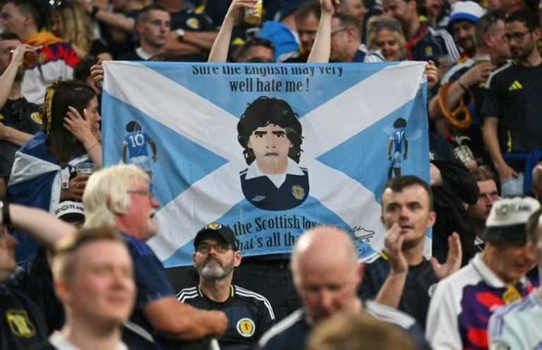 Crowd of football fans at a match holding a Scotland flag banner with a central caricature of a player and text along the top.