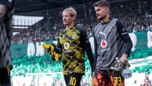 Two Borussia Dortmund goalkeepers in their kit walk off the pitch, each holding gloves as a crowded stadium cheers in the background.