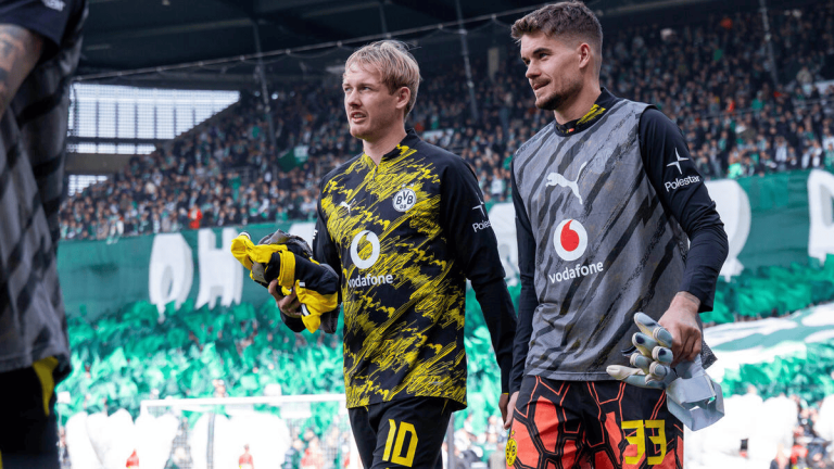 Two Borussia Dortmund goalkeepers in their kit walk off the pitch, each holding gloves as a crowded stadium cheers in the background.