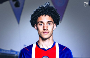 Portrait of a young man with curly hair wearing a blue and red soccer jersey, standing indoors with banners in the background.