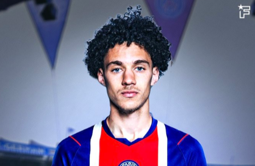 Portrait of a young man with curly hair wearing a blue and red soccer jersey, standing indoors with banners in the background.