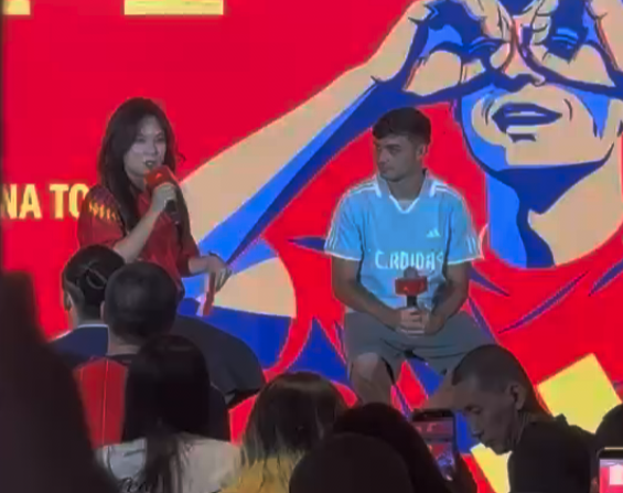 Woman in red speaks into a microphone on stage beside a man in a light blue jersey; bold red backdrop with graphics, audience in the foreground.
