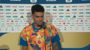 Colombian footballer at a press conference wearing a bright, multicolored jersey, speaking into a microphone with a FIFA World Cup backdrop behind him, sponsors visible.