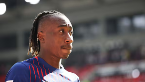 Male soccer player with braided hair in a blue kit, looking to the right on a blurred stadium field.