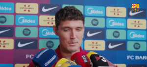 Footballer speaking at a press conference with a colorful sponsor backdrop and several microphones in front of him.