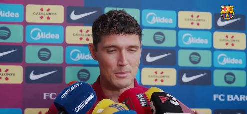 Footballer speaking at a press conference with a colorful sponsor backdrop and several microphones in front of him.
