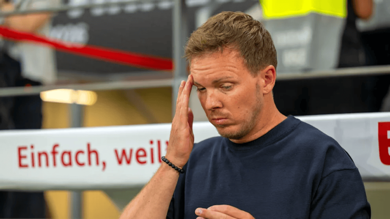 Man in a dark blue shirt touching his temple, looking stressed at an outdoor event with a red barrier in the background.