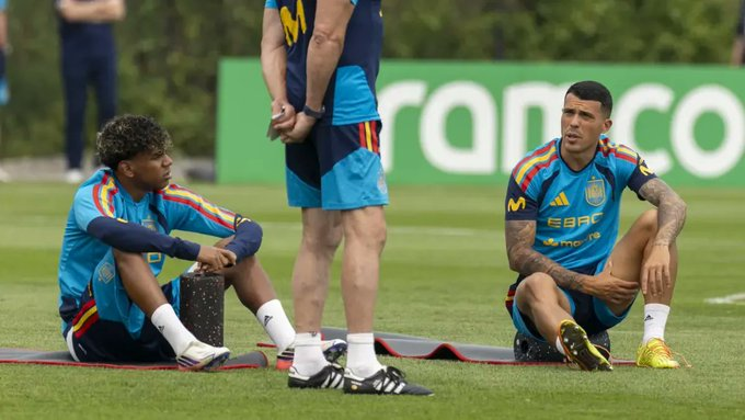 Two football players in blue training kits sit on the grass with red exercise mats during a practice session, while a teammate stands nearby; a green advertising board is visible in the background.