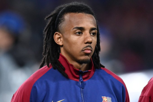 Male footballer wearing a blue and maroon FC Barcelona training jacket, looking thoughtful on the field.