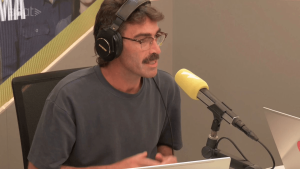 Man wearing headphones speaks into a yellow-matted mic in a radio studio, seated at a desk with a laptop nearby.