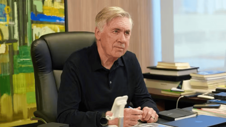 Older man seated at a desk in an office, looking thoughtful, with books stacked nearby and a colorful painting in the background.