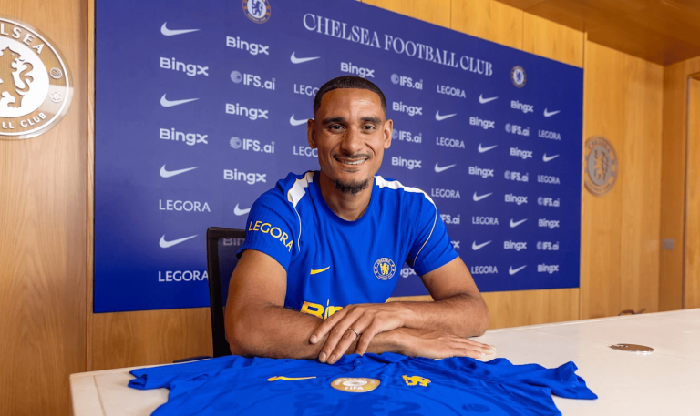 Chelsea footballer in a blue kit sits at a press conference table, smiling, with a sponsor-branded backdrop behind him and club crest nearby.
