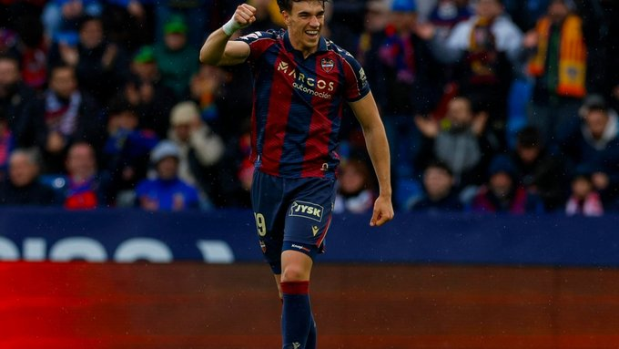 Soccer player in maroon and blue striped kit celebrates on the pitch with raised fist as fans cheer in the background, number 9 visible on shorts.