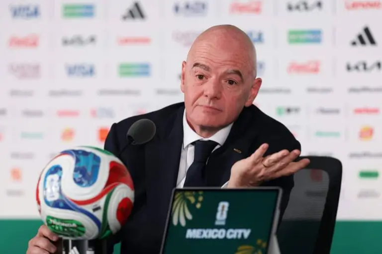 Man in a suit at a press conference with a soccer ball and a laptop displaying 'MEXICO CITY' logos in the foreground