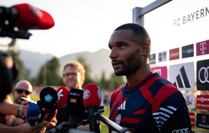 Male footballer in a red and blue training kit speaks to reporters during a post-match interview with multiple microphones in front of him.