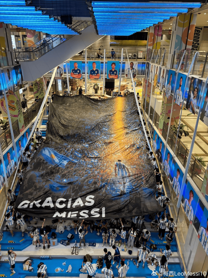 Large black banner reading 'GRACIAS MESSI' unfurled across a mall atrium by a crowd of Argentine fans beneath blue-lit ceilings.