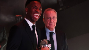 Two men in suits at an awards ceremony, the younger man laughing and holding a shiny spherical trophy as the older man smiles beside him