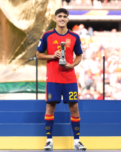 Footballer in Spain jersey stands on a podium, holding a trophy and smiling at an awards ceremony.