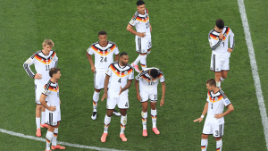 German national soccer team players standing in a circle on the field, looking down in white uniforms with black-red-yellow trim.