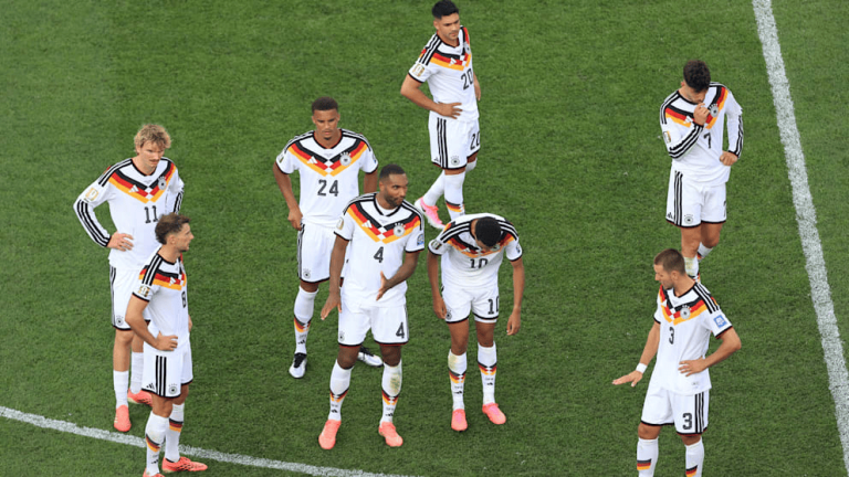 German national soccer team players standing in a circle on the field, looking down in white uniforms with black-red-yellow trim.