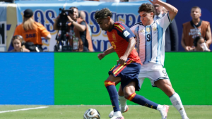 Two soccer players battle for the ball on a sunny field: one in a red jersey with blue shorts and the other in a light blue–white striped kit, near the sideline.