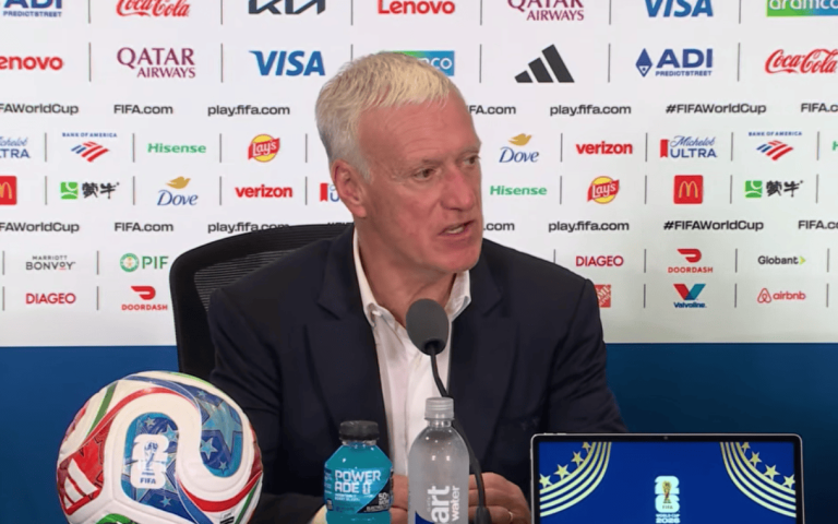 Man in a suit speaks at a FIFA press conference; sponsored backdrop, soccer ball and bottles on the table nearby.