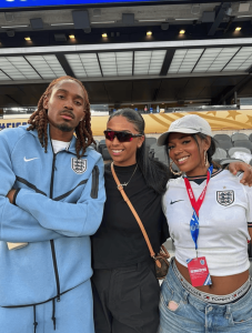 Three friends pose for a photo at a stadium; two wear England soccer jerseys (one in a light blue tracksuit, one in a white top with a cap), the third in black with sunglasses.