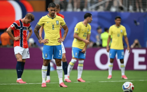 Brazilian players in yellow jerseys stand with hands on hips on a soccer field while a red-and-blue opponent looks down nearby; ball on the pitch ahead.
