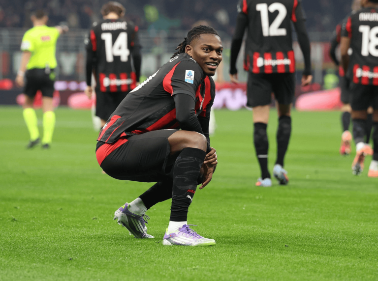 Soccer player in a black-and-red kit crouches on a grassy field during a match, looking toward the camera with teammates blurred in the background.