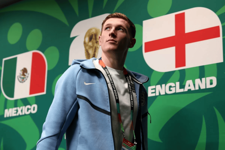 Young man in a light blue jacket standing before England and Mexico World Cup banners with a lanyard around his neck.