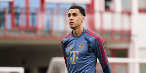 Young man in FC Bayern training kit runs on a field with a blurred red building behind.