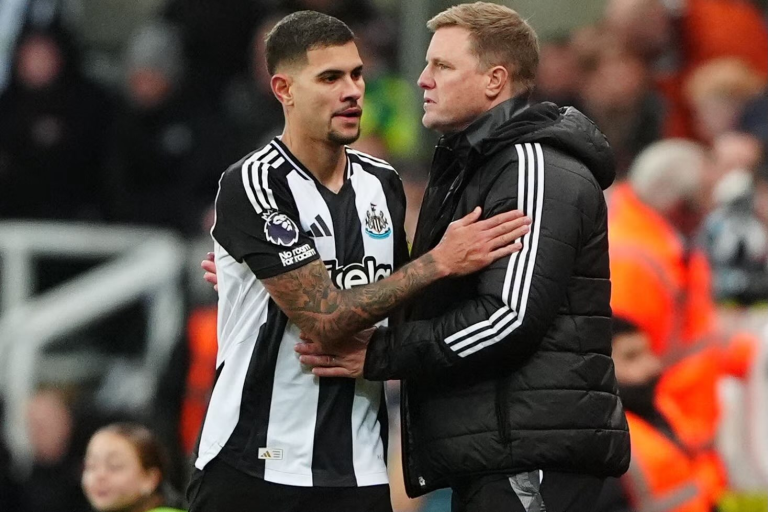 Soccer player in Newcastle United kit embraces a man in a black puffer jacket on the sideline.