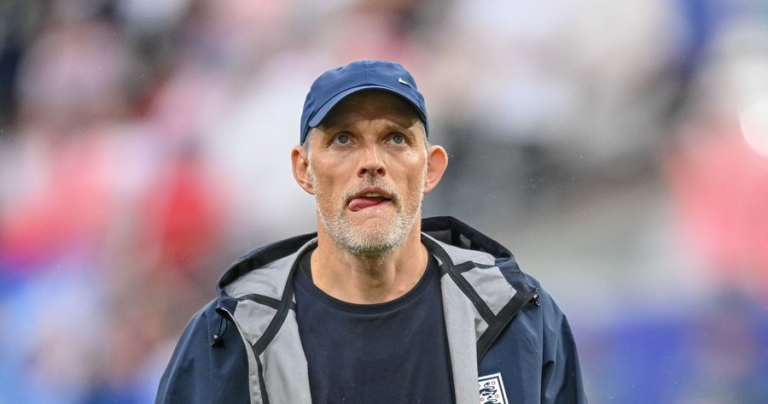Athletic man wearing a blue cap and navy jacket on a sports field, looking upward with a focused expression and his tongue slightly out; England crest visible on the jacket.
