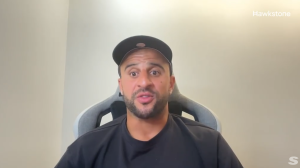 Man wearing a black t-shirt and cap sits in a gray gaming chair, facing the camera and speaking to the viewer.