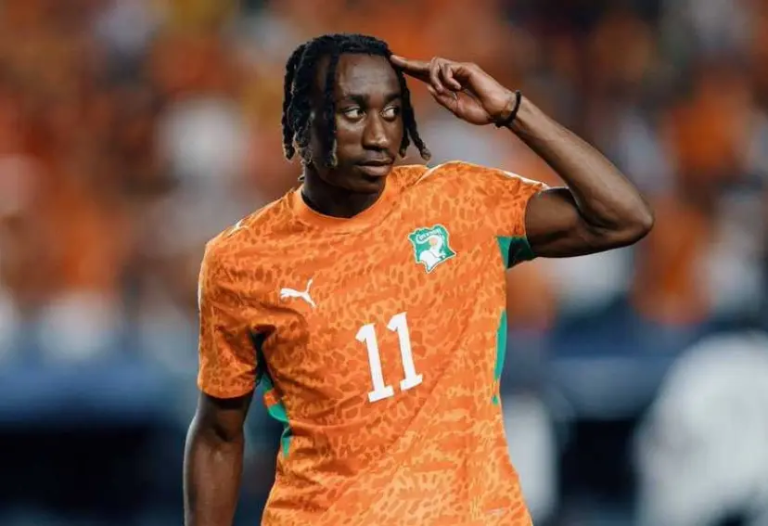 Soccer player in an orange Ivory Coast jersey with number 11 saluting with his right hand on a blurred stadium background.