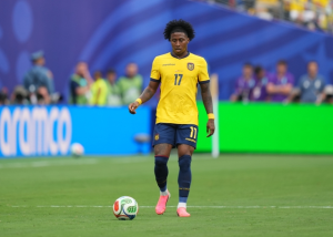 Soccer player in a yellow jersey with number 17 standing over a ball on a green pitch during a match with a blurred stadium background.