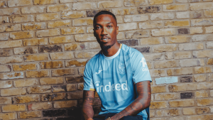 Man wearing a light blue soccer jersey sits against a brick wall, facing camera with a neutral expression, the jersey branded with 'indeed' and team crest visible.