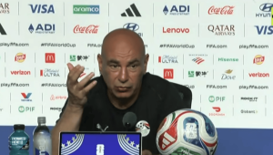 Bald man at a press conference gesturing with his hand, seated behind a table with a soccer ball and laptop screen in front of him.